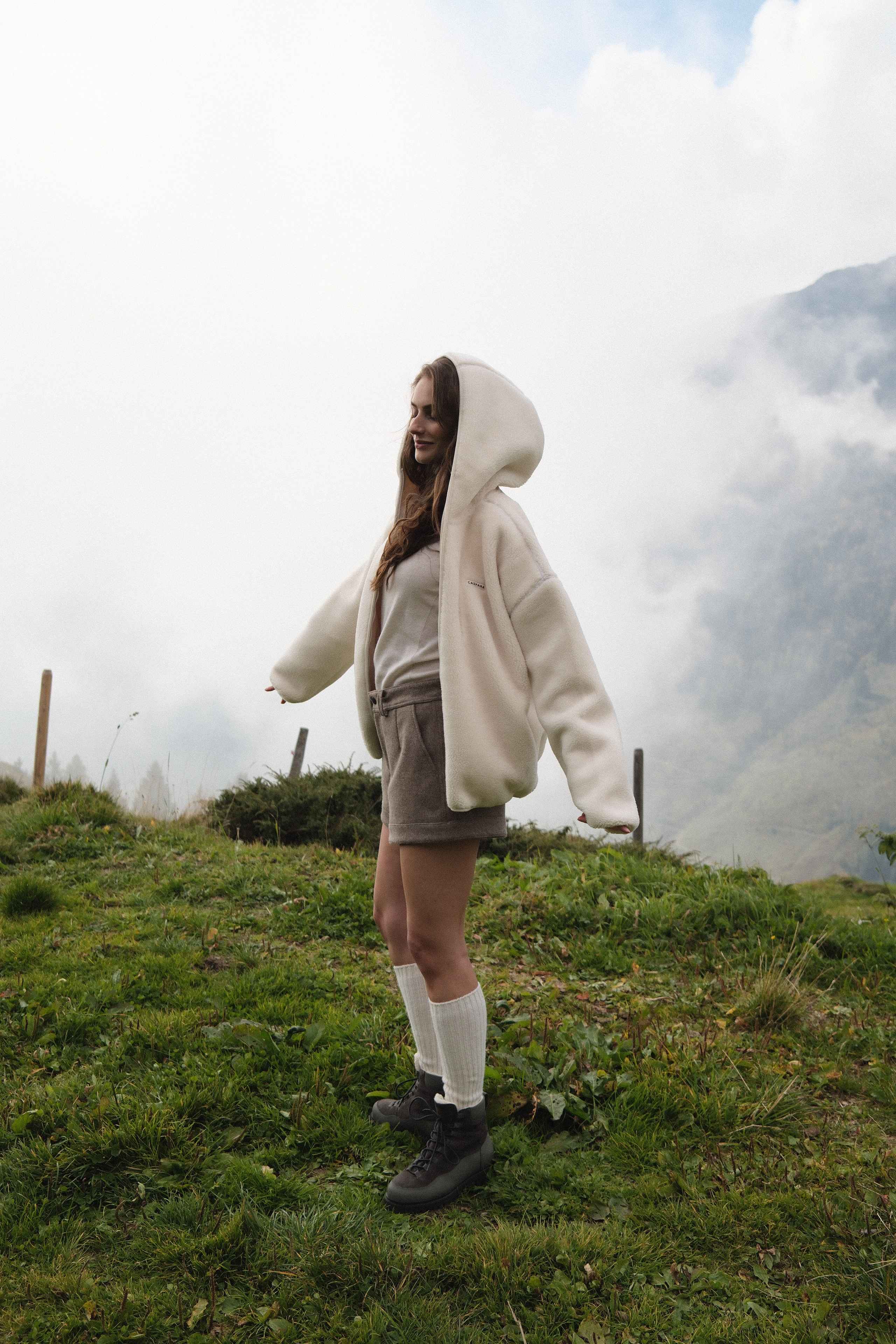 Woman wearing an oversized creme CASPARA hooded fleece jacket on a misty mountain slope.